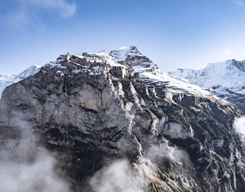 A majestic mountain range capped with snow under a clear blue sky. The rugged cliffs with layers of exposed rock create a dramatic and breathtaking landscape. Mist floats gently around the base, adding a mystical touch to the scene.