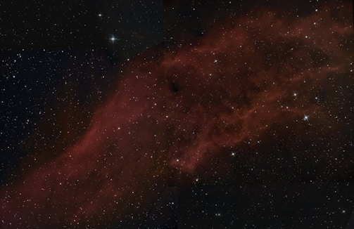 A vivid display of a nebula with a rich red hue stretching diagonally across the image, surrounded by a vast multitude of twinkling stars against a dark backdrop.