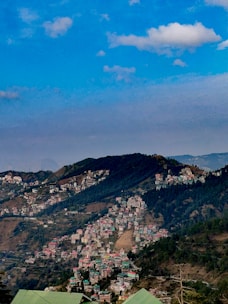 A scenic hill station with numerous houses clustered on the slopes of lush green mountains under a blue sky with scattered clouds.