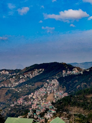 A scenic hill station with numerous houses clustered on the slopes of lush green mountains under a blue sky with scattered clouds.