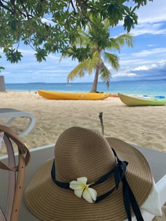A tropical beach setting features a sandy shore with a couple of kayaks resting on it, one yellow and the other light green. In the foreground, a straw hat adorned with a white flower and black ribbon sits on a table, partially shadowed by overhanging greenery. A palm tree stands prominently in the background near the turquoise ocean under a clear blue sky.