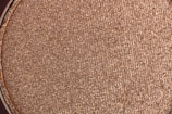 A close-up view of a textured, shimmering brown surface. The material appears to be finely powdered with a metallic sheen, suggesting it could be makeup or a similar product.