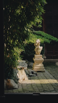 A serene garden scene featuring a stone sculpture on a pedestal surrounded by lush green foliage. The pathway is cobblestone, leading toward a traditional structure with intricate wooden architecture.