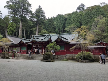 A traditional Japanese temple with red wooden structures and intricate roof designs surrounded by lush green forest. Several people are present at the entrance, and the temple sits on a gravel courtyard with scattered vegetation and stone sculptures.