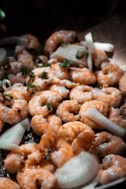 A close-up of shrimp being sautéed with slices of onion and garnished with green herbs in a pan. The shrimp are cooked to a golden brown, glistening with a layer of oil.