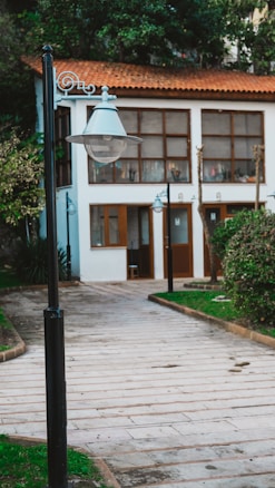 A paved walkway lined with greenery leads to a quaint white building with wooden doors and windows. A classic street lamp is prominently featured in the foreground, and the building has a warm red-tiled roof. The setting appears peaceful and well-maintained.