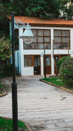 A paved walkway lined with greenery leads to a quaint white building with wooden doors and windows. A classic street lamp is prominently featured in the foreground, and the building has a warm red-tiled roof. The setting appears peaceful and well-maintained.