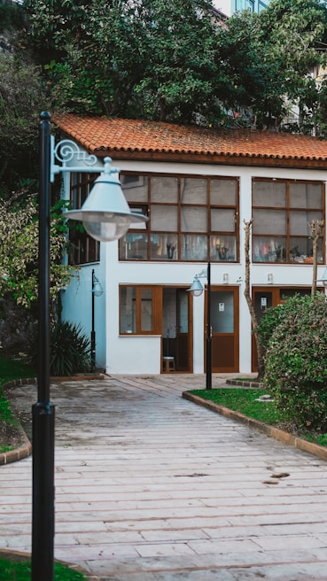 A charming path lined with street lamps leads to a small building with a terracotta roof and large windows. The building is nestled among lush greenery, including bushes and trees, which add a natural touch to the setting.
