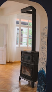A cozy room with a traditional black wood stove stands against a white wall with blue artistic swirls. The room features wooden parquet flooring and white curtains that allow natural light through a set of tall windows.