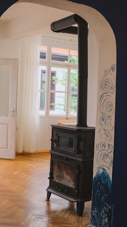 A cozy room with a traditional black wood stove stands against a white wall with blue artistic swirls. The room features wooden parquet flooring and white curtains that allow natural light through a set of tall windows.