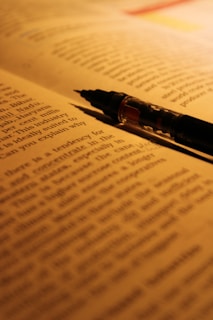 A close-up view of a book with a pen placed diagonally on top of the open pages. The text is in focus at the top and gradually becomes blurred towards the bottom of the page. Warm lighting creates a cozy and intimate atmosphere.
