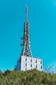 A tall communication tower rises from a modern industrial building, equipped with antennas and satellite dishes. The structure is surrounded by green foliage and set against a clear blue sky.