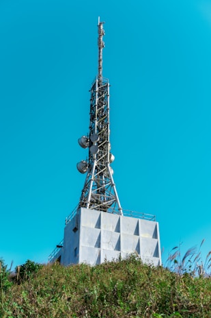 A tall communication tower rises from a modern industrial building, equipped with antennas and satellite dishes. The structure is surrounded by green foliage and set against a clear blue sky.