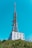 A tall communication tower rises from a modern industrial building, equipped with antennas and satellite dishes. The structure is surrounded by green foliage and set against a clear blue sky.