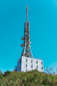 A tall communication tower rises from a modern industrial building, equipped with antennas and satellite dishes. The structure is surrounded by green foliage and set against a clear blue sky.