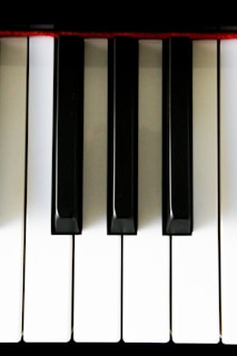 Close-up view of piano keys with a pattern of alternating black and white keys. The image is well-lit, highlighting the contrast between colors. A hint of red fabric is visible on the upper edge.