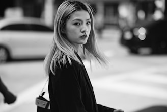 A black and white photo of a young woman with long, light hair standing in a city setting. She is wearing a dark coat and looking over her shoulder with a thoughtful expression. Blurred cars and buildings can be seen in the background, creating a sense of urban motion.
