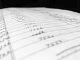 A close-up view of a handwritten page filled with numbers and words organized into columns and rows. The paper appears slightly tilted, with some entries in sharp focus while others blur in the background, suggesting depth and perspective.