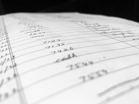 A close-up view of a handwritten page filled with numbers and words organized into columns and rows. The paper appears slightly tilted, with some entries in sharp focus while others blur in the background, suggesting depth and perspective.