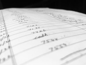A close-up view of a handwritten page filled with numbers and words organized into columns and rows. The paper appears slightly tilted, with some entries in sharp focus while others blur in the background, suggesting depth and perspective.