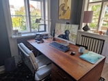 A well-organized home office with a wooden desk holding a monitor displaying a dog, keyboard, mouse, and a mug. An office chair is placed in front of the desk. On the desk, there is also an old-fashioned rotary phone, a lamp, and some headphones. The room has two large windows providing ample natural light. A painting hangs on the wall and various items, including a laptop and documents, are present on the desk and window sills, creating a cozy workspace environment.