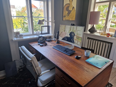 A well-organized home office with a wooden desk holding a monitor displaying a dog, keyboard, mouse, and a mug. An office chair is placed in front of the desk. On the desk, there is also an old-fashioned rotary phone, a lamp, and some headphones. The room has two large windows providing ample natural light. A painting hangs on the wall and various items, including a laptop and documents, are present on the desk and window sills, creating a cozy workspace environment.