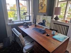 A well-organized home office with a wooden desk holding a monitor displaying a dog, keyboard, mouse, and a mug. An office chair is placed in front of the desk. On the desk, there is also an old-fashioned rotary phone, a lamp, and some headphones. The room has two large windows providing ample natural light. A painting hangs on the wall and various items, including a laptop and documents, are present on the desk and window sills, creating a cozy workspace environment.