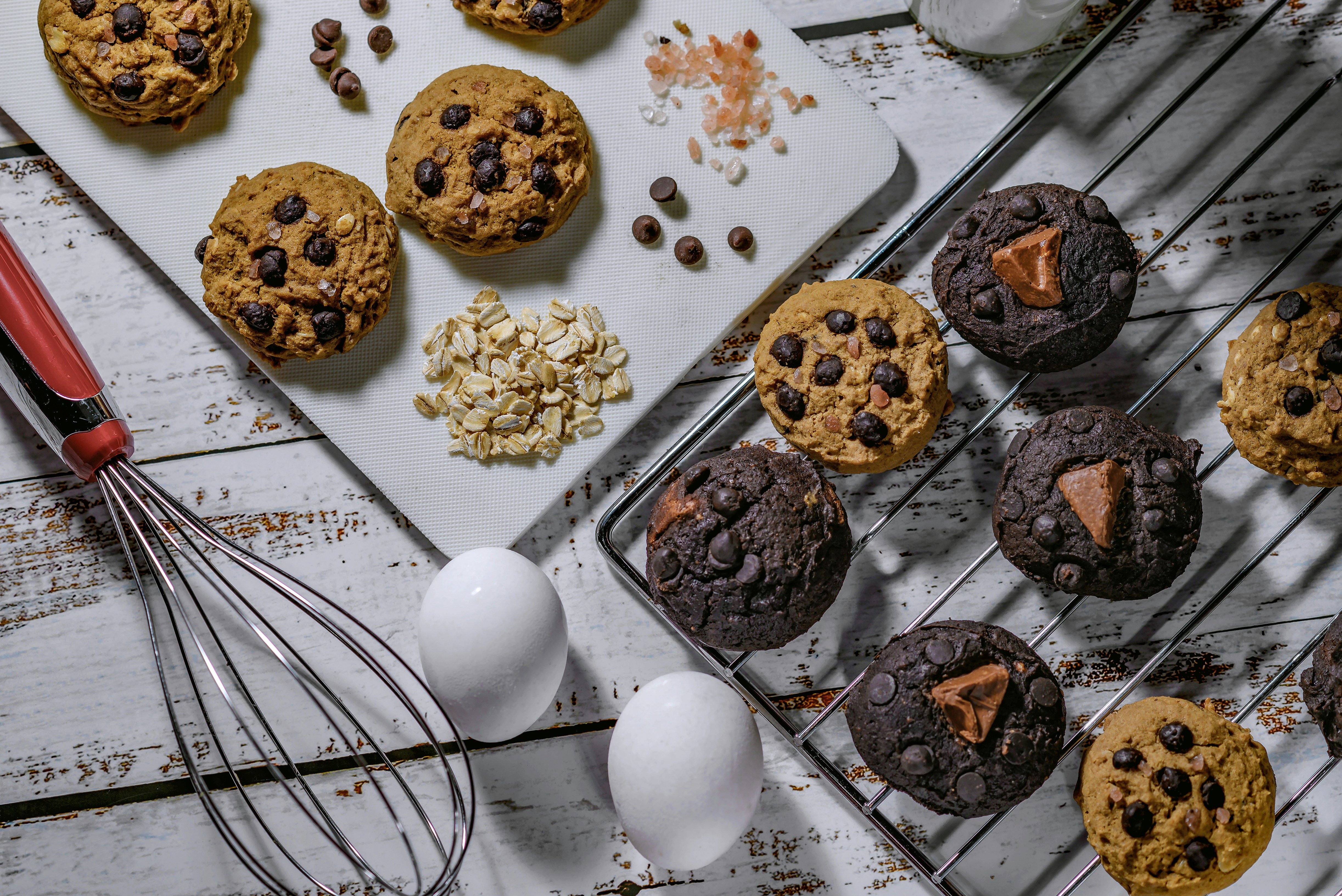 A selection of homemade cookies arranged on a white wooden surface, with some placed on a cooling rack and others on a cutting board. The cookies are dotted with chocolate chips and some have chunks of chocolate. Near the cookies are ingredients including rolled oats, chocolate chips, and Himalayan pink salt. A whisk with a red handle, two white eggs, and a glass bottle of milk are also present.