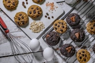A selection of homemade cookies arranged on a white wooden surface, with some placed on a cooling rack and others on a cutting board. The cookies are dotted with chocolate chips and some have chunks of chocolate. Near the cookies are ingredients including rolled oats, chocolate chips, and Himalayan pink salt. A whisk with a red handle, two white eggs, and a glass bottle of milk are also present.