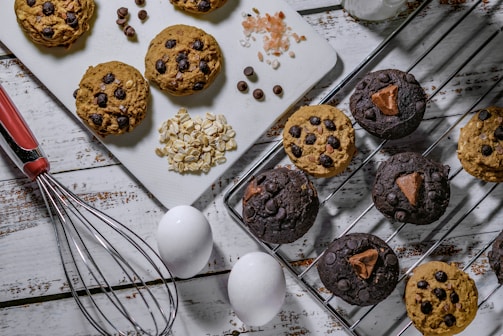 A selection of homemade cookies arranged on a white wooden surface, with some placed on a cooling rack and others on a cutting board. The cookies are dotted with chocolate chips and some have chunks of chocolate. Near the cookies are ingredients including rolled oats, chocolate chips, and Himalayan pink salt. A whisk with a red handle, two white eggs, and a glass bottle of milk are also present.
