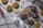 A selection of homemade cookies arranged on a white wooden surface, with some placed on a cooling rack and others on a cutting board. The cookies are dotted with chocolate chips and some have chunks of chocolate. Near the cookies are ingredients including rolled oats, chocolate chips, and Himalayan pink salt. A whisk with a red handle, two white eggs, and a glass bottle of milk are also present.