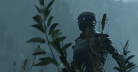 A robotic figure with glowing eyes is positioned among tall plants in a foggy, natural setting, evoking a sense of mystery and technology blended with nature.
