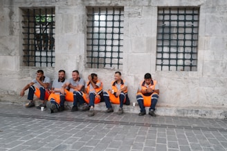 A group of six construction workers wearing orange safety vests sits on a sidewalk against an old stone wall with barred windows. They appear to be taking a break, with some drinking from cups and others using their phones. The ground is paved with stone tiles.