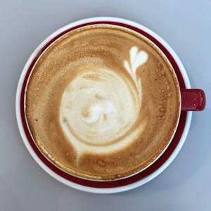 A close-up view of a beautifully crafted latte with a swirling latte art design in a red cup with a white saucer against a light gray background.