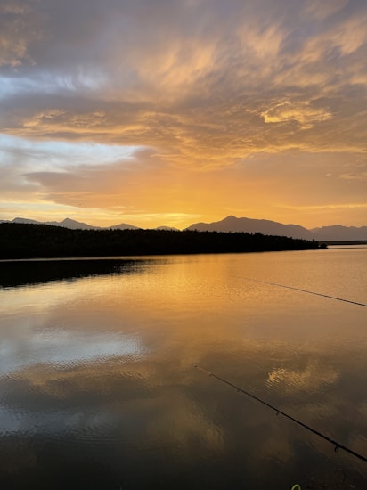 A serene lake reflects the vibrant colors of a sunset, with hues of orange and yellow dominating the sky. Mountains silhouette against the sky in the background, and a fishing rod subtly extends into the water from the bottom right corner.