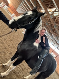 A person with blonde hair and a tattoo on their arm is joyfully hugging a large black and white horse in an indoor riding arena. The arena is spacious with a high wooden ceiling and sand-covered ground. The horse is wearing a bridle, and there is another person partially visible holding its reins.