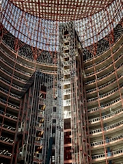 An interior view of a high-rise building with a striking architectural design. The structure is circular with numerous floors visible, each lined with glass and metal frameworks. The ceiling is a geometric grid of metal beams, allowing light to filter through. The use of red and metallic colors gives the building a modern and industrial appearance.