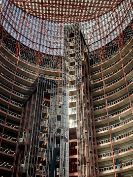An interior view of a high-rise building with a striking architectural design. The structure is circular with numerous floors visible, each lined with glass and metal frameworks. The ceiling is a geometric grid of metal beams, allowing light to filter through. The use of red and metallic colors gives the building a modern and industrial appearance.