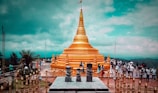 A large golden stupa dominates the scene, set against a backdrop of lush greenery and a partly cloudy sky. Numerous people are walking around the stupa, some dressed in traditional clothing. The structure is surrounded by ornate golden railings and stone sculptures, adding to the cultural significance of the location.