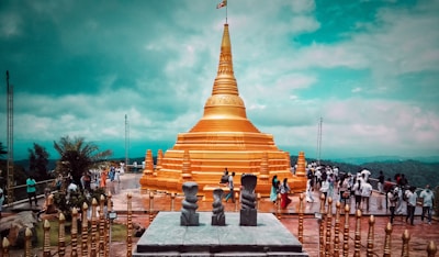 A large golden stupa dominates the scene, set against a backdrop of lush greenery and a partly cloudy sky. Numerous people are walking around the stupa, some dressed in traditional clothing. The structure is surrounded by ornate golden railings and stone sculptures, adding to the cultural significance of the location.