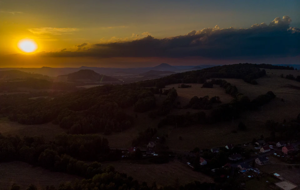 The image presents an aerial view of a rural landscape, with a large body of water and rolling hills in the background. The sun is setting behind the hills, casting a warm glow over the scene. The sky is filled with clouds, adding depth to the image. In the foreground, there are several houses scattered across the landscape, suggesting a small community living off-grid.