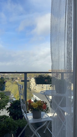 A small balcony scene with a metal table and chairs, decorated with a variety of potted plants and flowers. A sheer curtain is draped to the right, partially obscuring the view. The backdrop includes a cityscape with buildings, trees, and a partly cloudy sky.