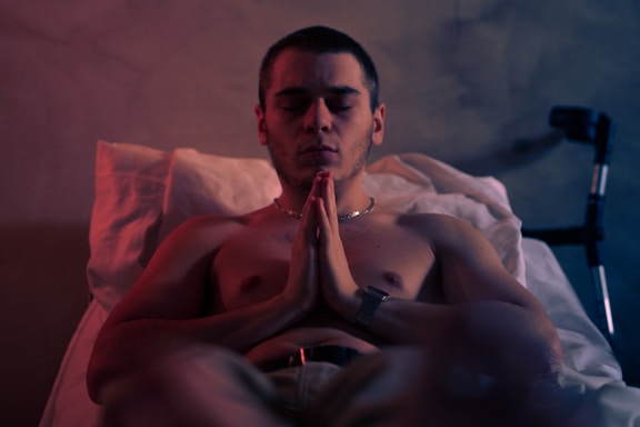 A shirtless man is seated with his hands pressed together in a prayer-like posture. He appears to be meditating or deeply focused, with closed eyes and slightly furrowed brows. The setting includes a bed with white sheets and a walking cane leaning in the background. There is soft ambient lighting with shades of red and blue, casting gentle shadows.