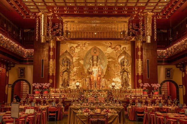 A richly decorated religious hall featuring a large, ornate altar with three golden statues in the center. The statues are surrounded by intricate woodwork and detailed carvings with a backdrop depicting celestial scenes. Tables with elaborate settings and floral arrangements fill the foreground, while the ceiling displays an ornate pattern and hanging lanterns.