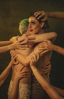 A dramatic scene where two individuals embrace each other closely, surrounded by multiple arms reaching towards them. One person has notable green hair and is primarily turned away, while the other person has striking makeup with bold eyeliner and is looking to the side. The background is dark, emphasizing the emotional intensity of the scene.