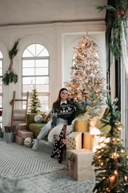 A cozy, elegantly decorated holiday room featuring a woman sitting on a green sofa. She wears a festive sweater with a winter scene and holds her hand to her cheek. A beautifully adorned Christmas tree with white and red ornaments stands beside her, surrounded by wrapped presents. Soft, warm lighting enhances the atmosphere, with additional greenery and decorative lights completing the scene.