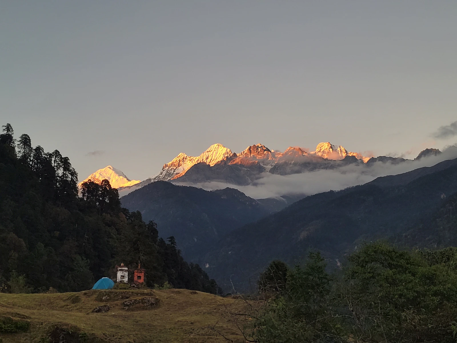 Low sun turning an arc of Kanchenjunga range summits gold above dark forested ridges, two chortens and a tent in a trek camp meadow