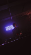 A DJ is performing in a dimly lit environment with a prominent purple neon sign in the background that reads 'VERTU'. The atmosphere is dark with subtle lighting focused on the DJ, who is wearing headphones and working behind a console.