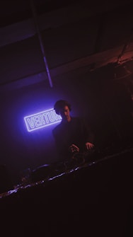 A DJ is performing in a dimly lit environment with a prominent purple neon sign in the background that reads 'VERTU'. The atmosphere is dark with subtle lighting focused on the DJ, who is wearing headphones and working behind a console.