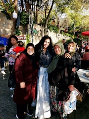 A group of women standing together outdoors, surrounded by trees and people in a lively environment. Some are wearing headscarves and smiling, while others are engaged in conversations. Various people and elements like trees, a stone wall, and a red canopy add depth to the setting.